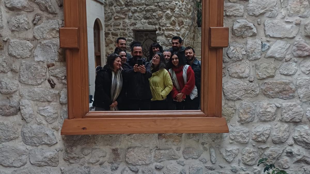 Tahta çerçeveli aynada yansıyan sekiz kişilik grup, taş duvar, beyaz masa ve sandalyeler.