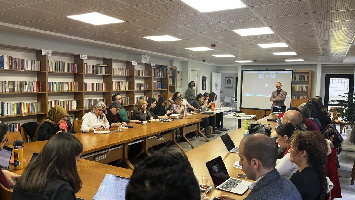 Konferans odasında uzun masalarda toplantı yapan insanlar, sunum izliyor.