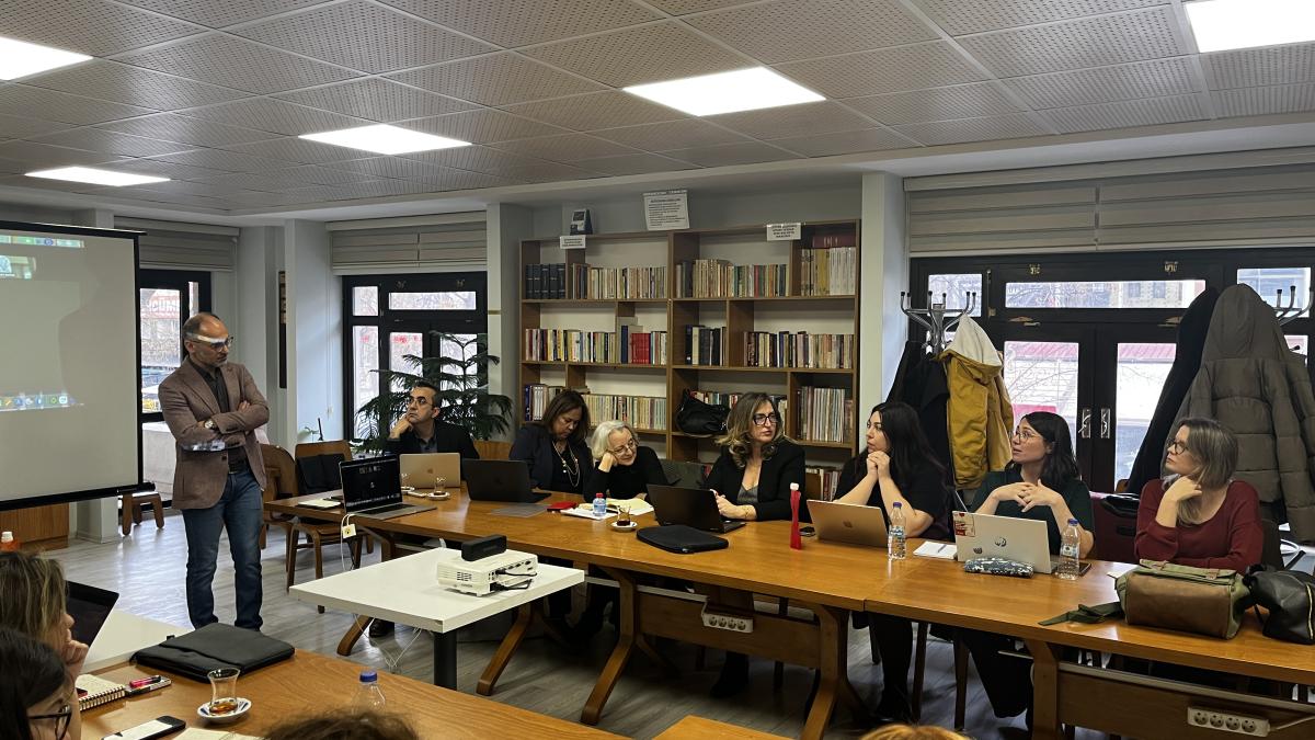 Bir sınıfta toplantı yapan insanlar, projeksiyon ekranı ve kitap rafları arka planda.