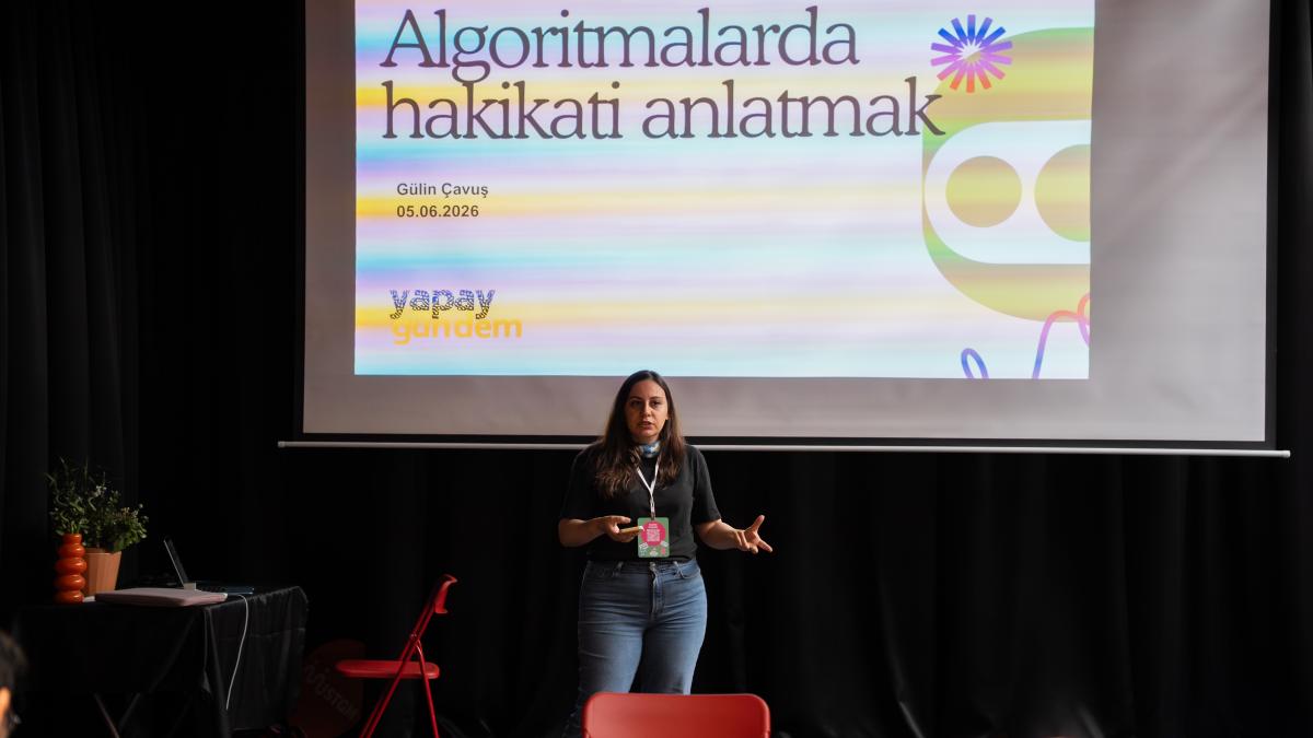 Bir sahnede sunum yapan kadın, arka planda "Algoritmalarda hakikati anlatmak" yazılı slayt.