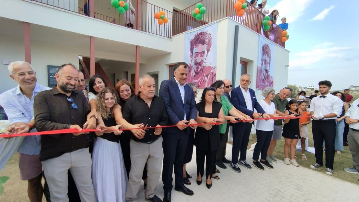Bir grup insan, açılışta kırmızı kurdele kesiyor; arkada balonlar ve büyük portreler var.