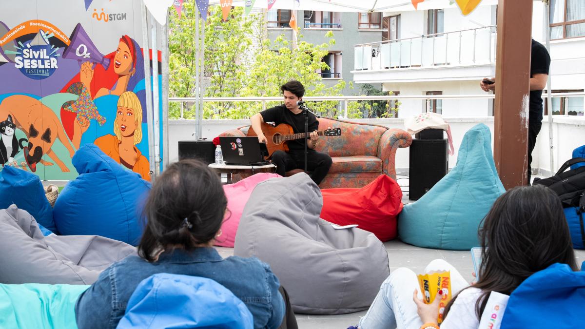 Açık havada puflarda oturan kişiler, gitar çalan bir müzisyeni dinliyor.
