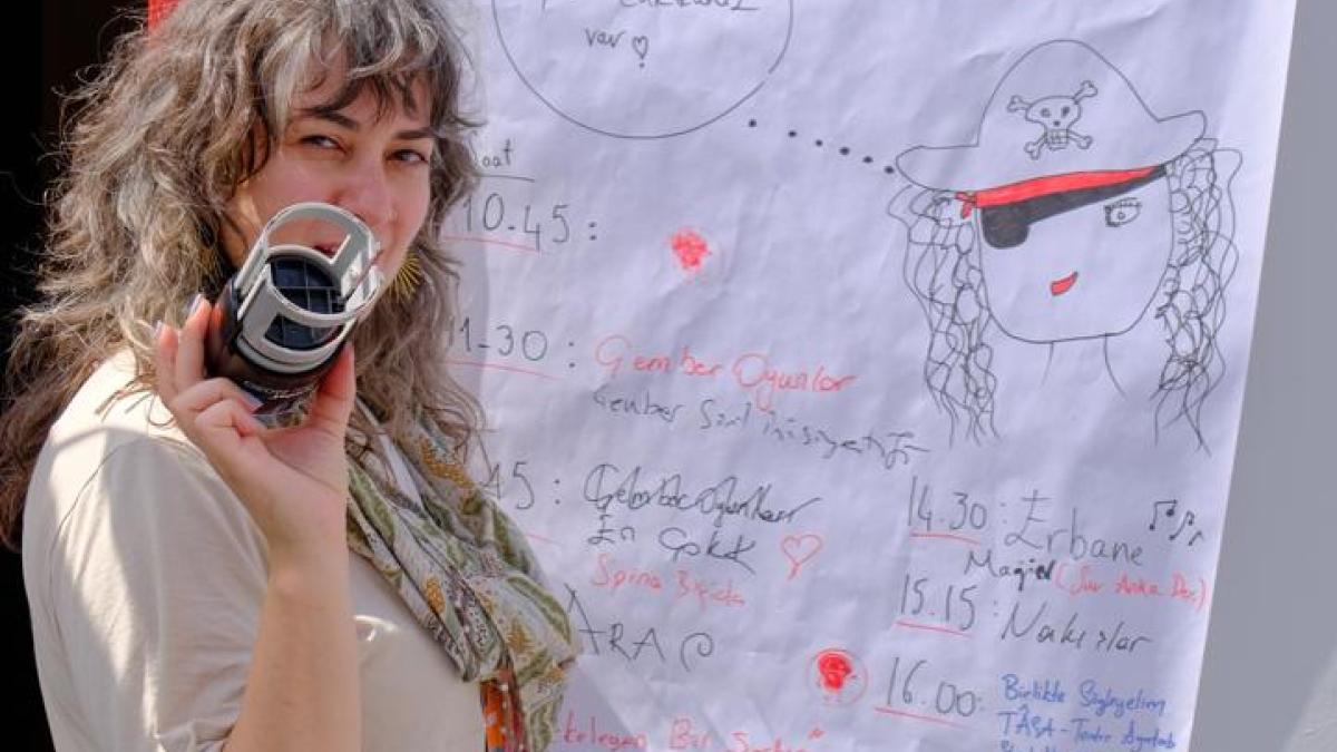 Elinde kamera tutan kadın, arkasında duvarda büyük bir çizim posteri var.
