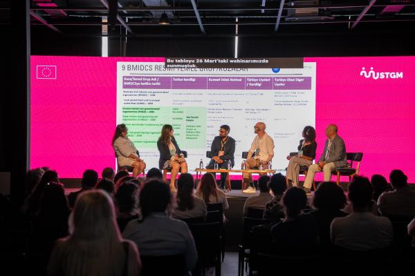 Pembe arka planda sahnede altı kişi panelde konuşuyor, izleyiciler önde oturuyor.