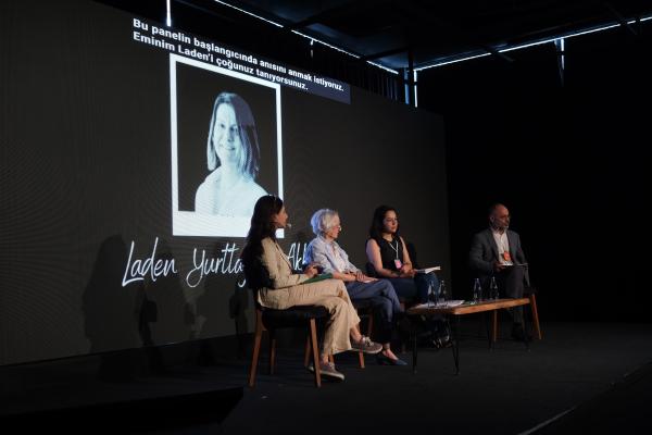 Sahnede dört panelist ve arkada büyük bir ekran, ekranda bir kadın fotoğrafı.