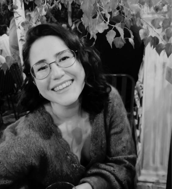Gülümseyen genç kadın, gözlüklü ve dalgalı saçlı, yapraklı bir ortamda oturuyor.