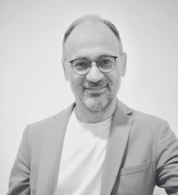 Gözlüklü, sakallı adam takım ceket ve tişörtle gülümsüyor.